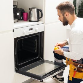 repairman examining oven with screwdriver in kitchen with tool case